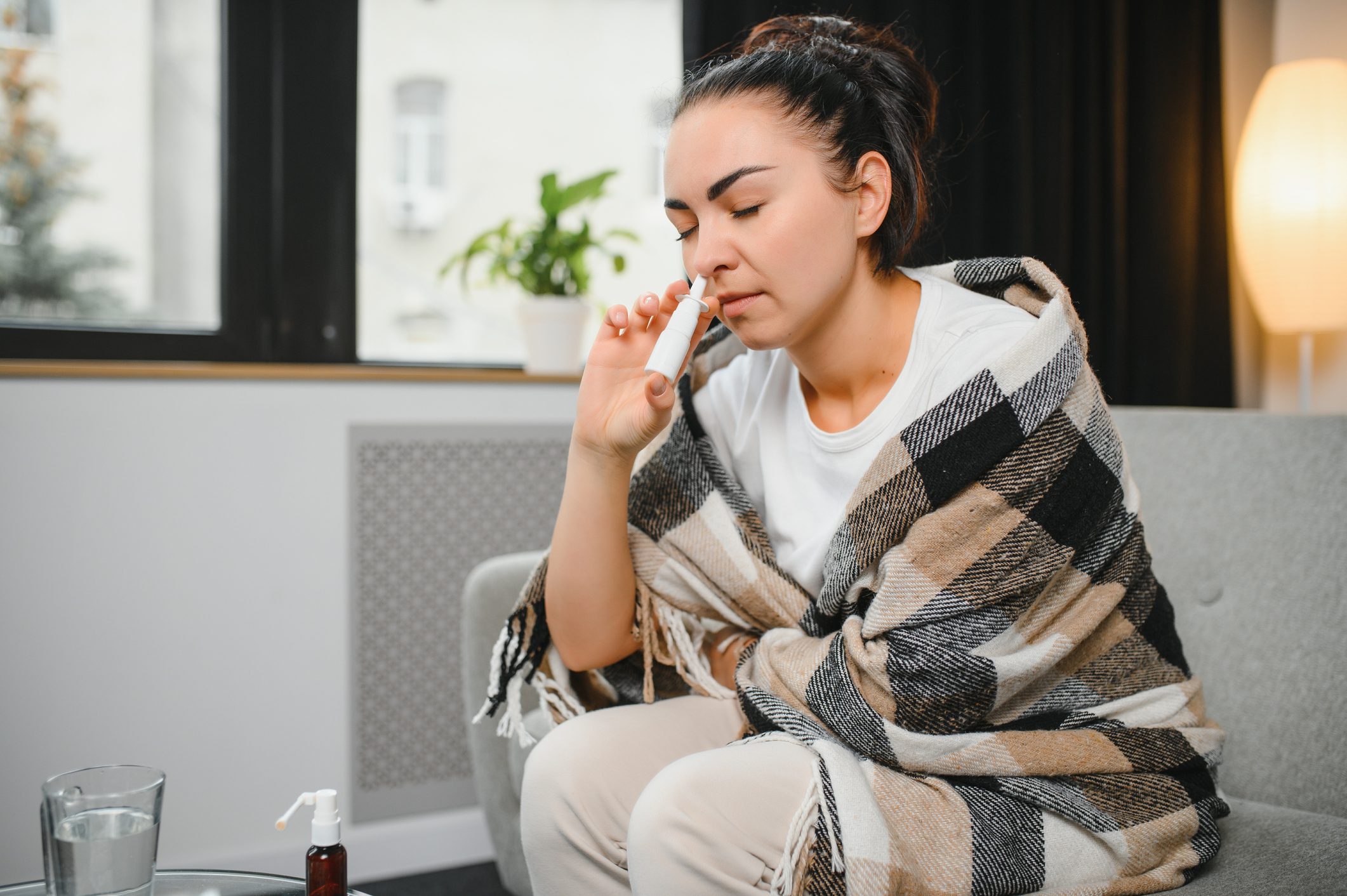 Nasal spray in hand of sick young woman sitting on couch. Allergic rhinitis symptoms and treatment