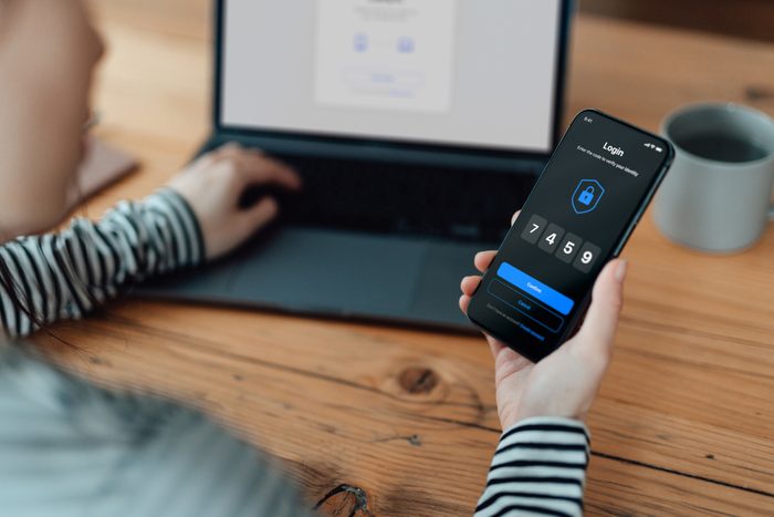 Over the shoulder view of young woman using mobile app with Two-Factor Authentication (2FA) security system to access online bank account via laptop