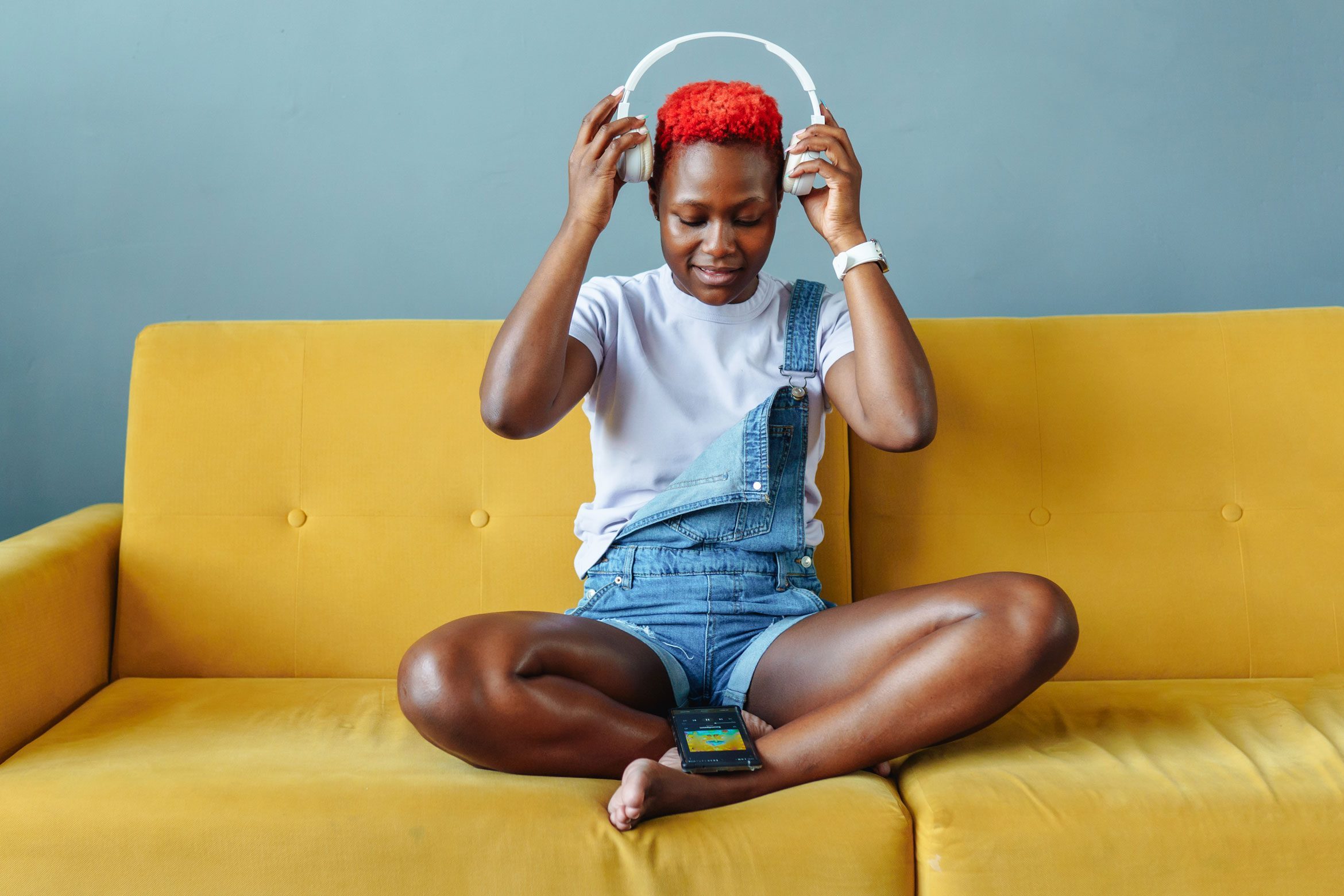 Young woman putting on headphones, sitting on sofa in lotus pose