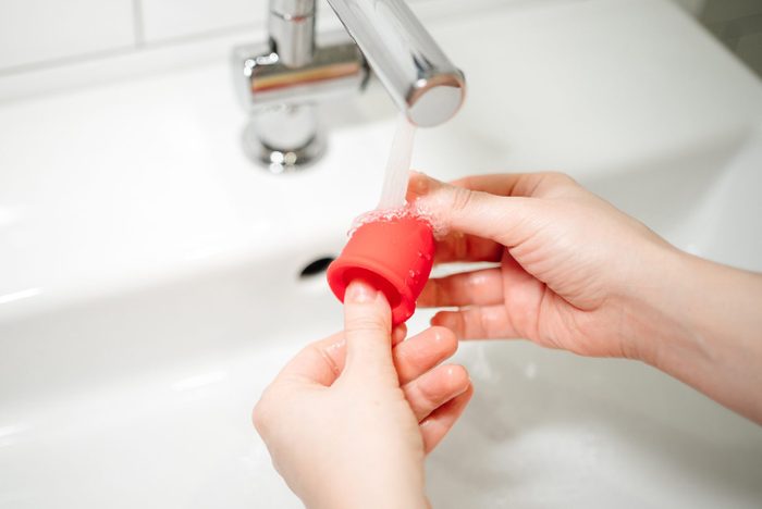 Hands of a woman washes a red menstrual cup with water under the tap at the sink. Alternative feminine hygiene product concept during menstruation.