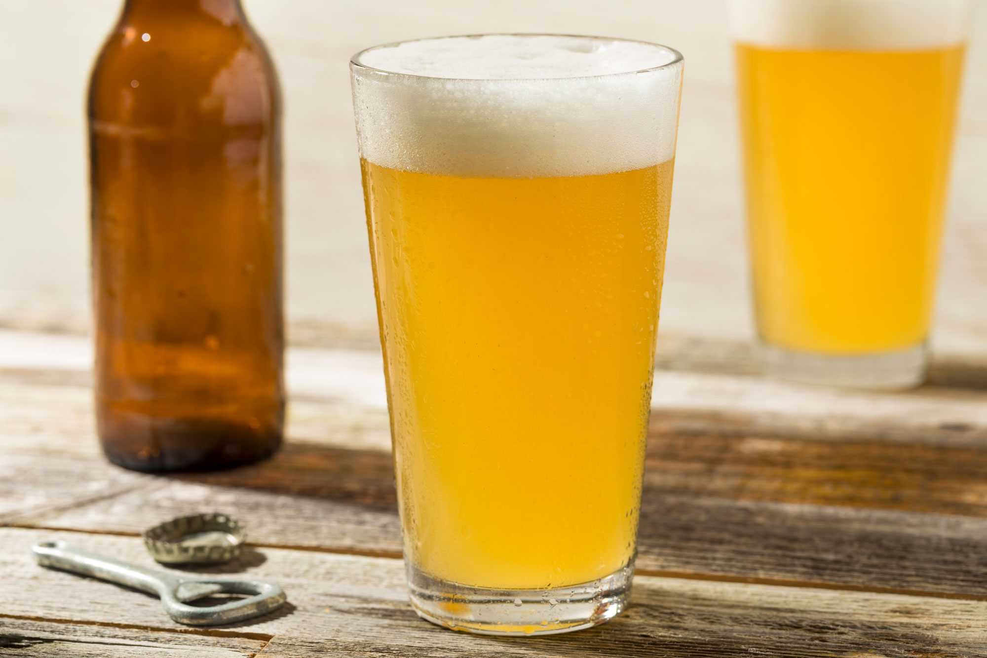 light beer in a glass on wood surface with bottle opener, bottle, and second glass in the background