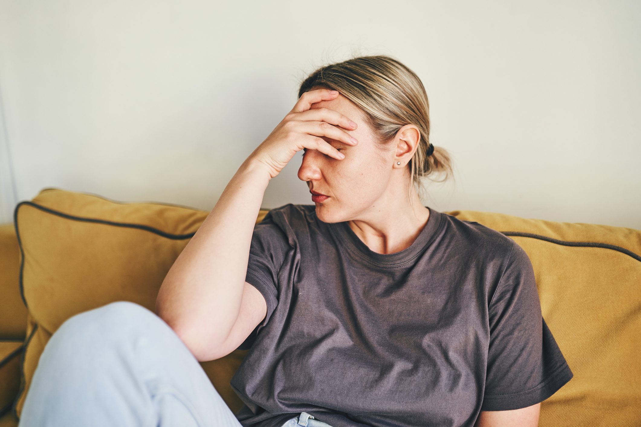 A young woman is sitting on the yellow couch at home with her head in her hands