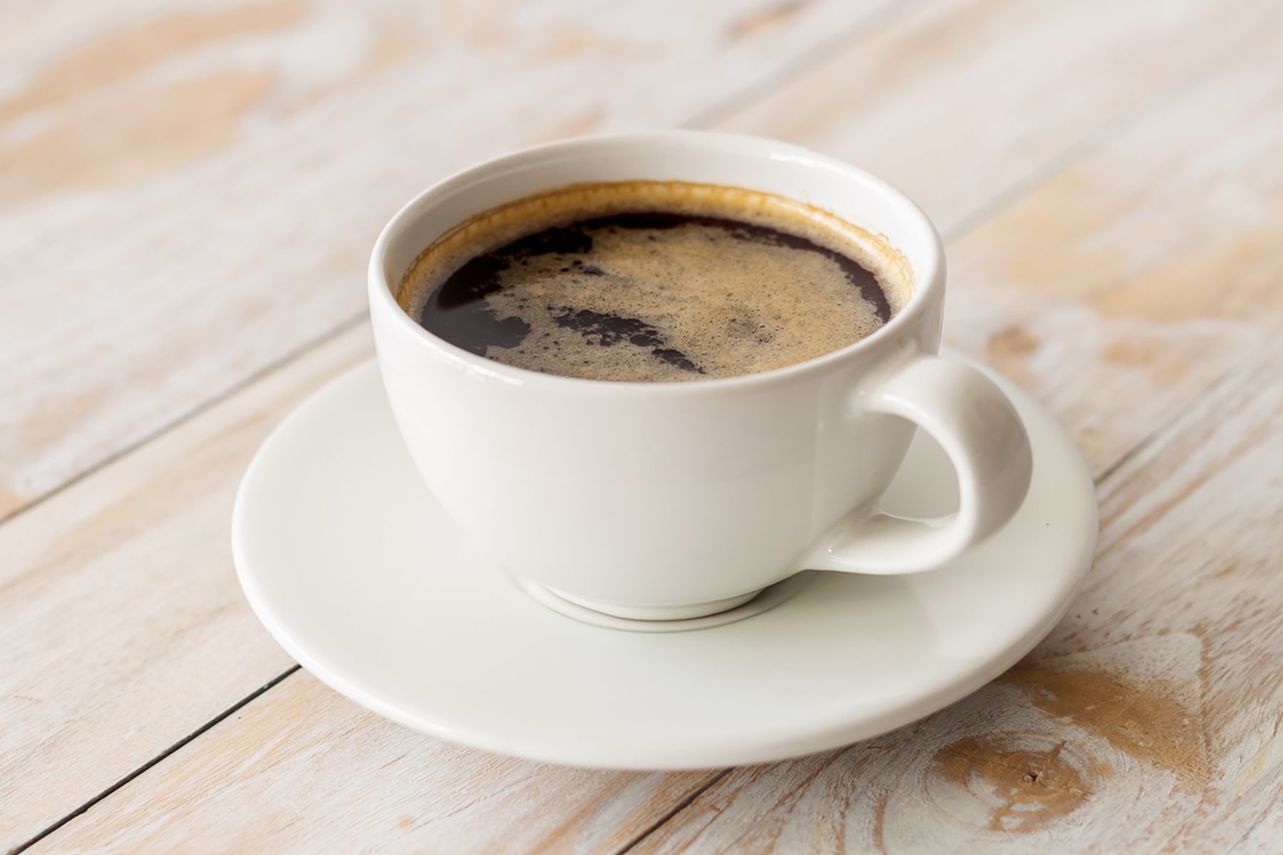 A white ceramic cup filled with black coffee sits on a matching white saucer. The coffee has a light frothy layer on top, on the light wooden table next to it.