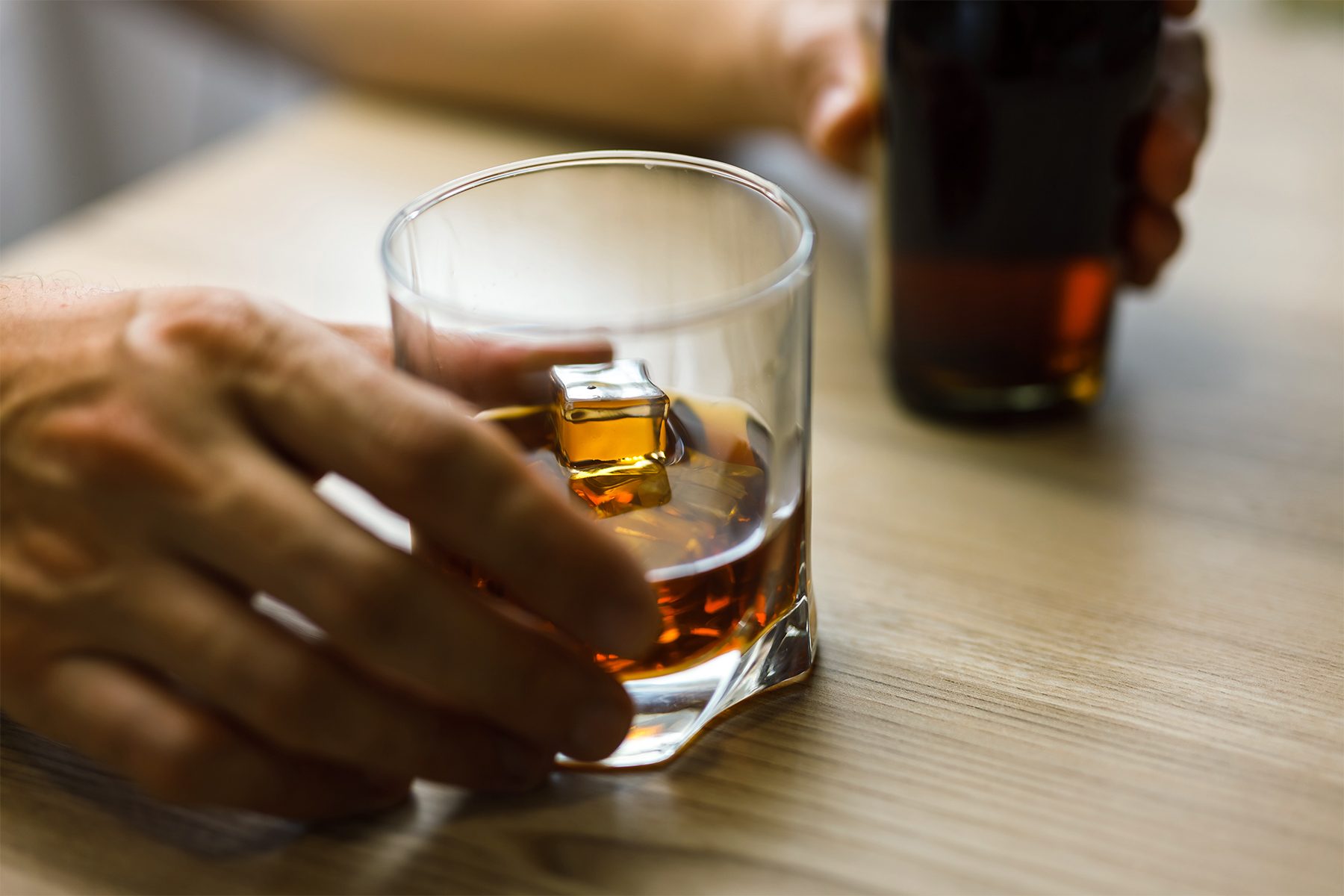 A person holding a glass of whiskey with a hand visible, while another hand holds a bottle on a wooden table. The liquid in the glass is amber-colored.