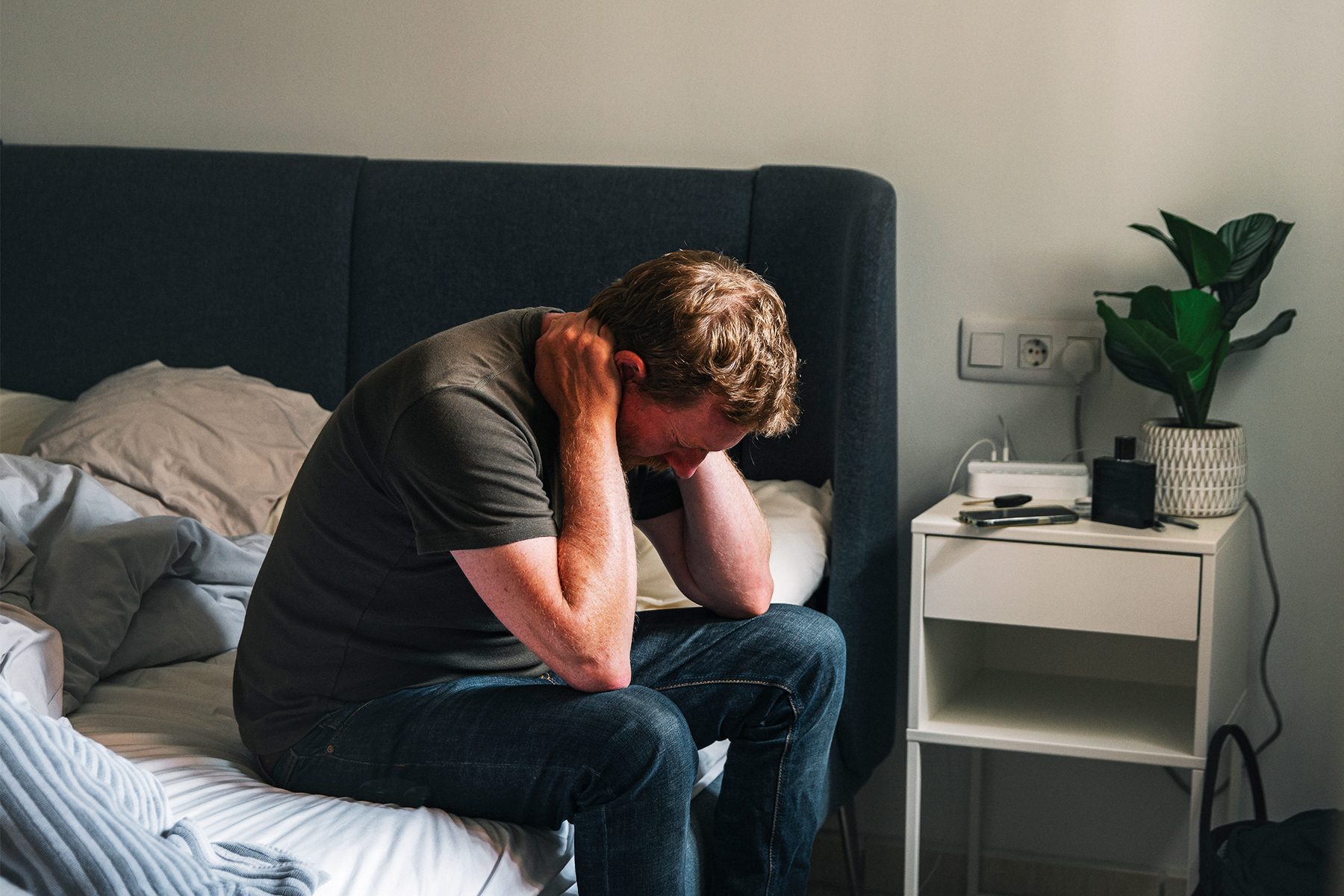 A depressed mature male sits on his bed. He clasps his hand behind his head