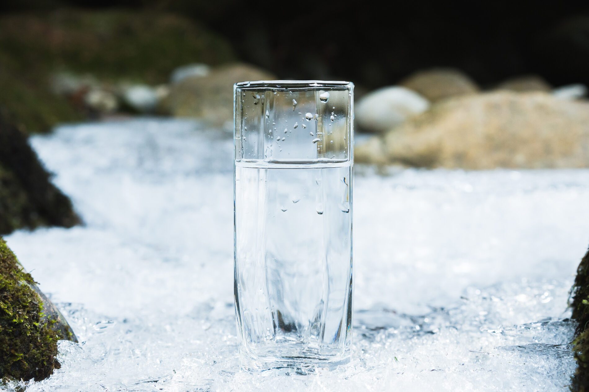 Transparent glass glass with drinking mountain water in winter stands on an icy crust against the background of a clean, frosty river bank. The concept of drinking mountain drinking mineral water and the production of mineral ecologically clean drinking