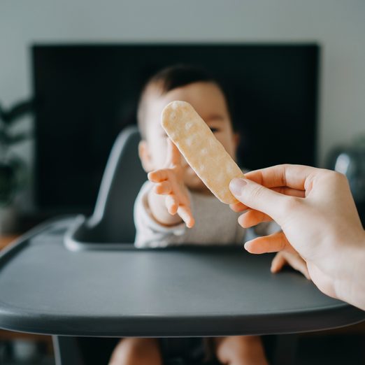 Hungry Baby Reaching For Finger Biscuit From Mother's Hand On High Chair